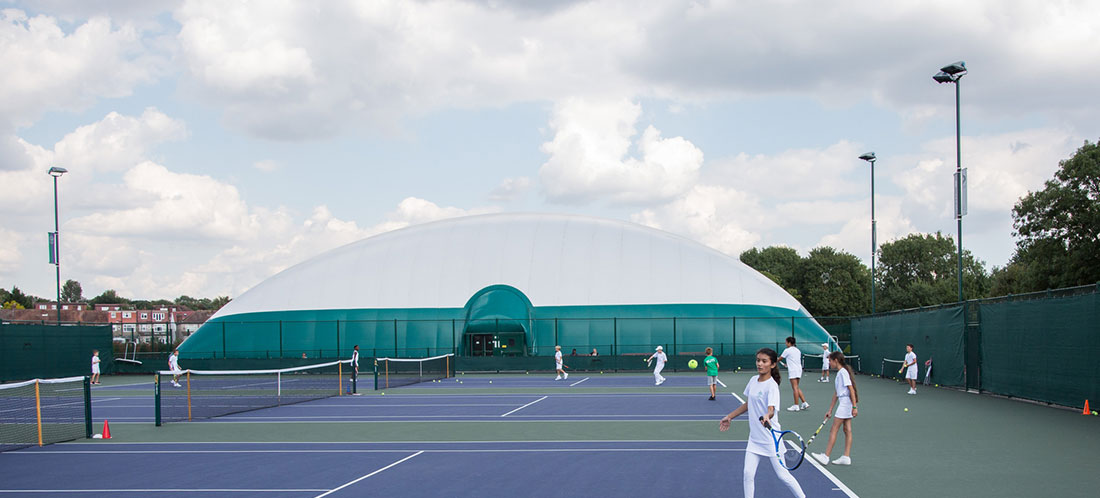 Children play tennis on 3 tennis courts in front of a large green and white dome